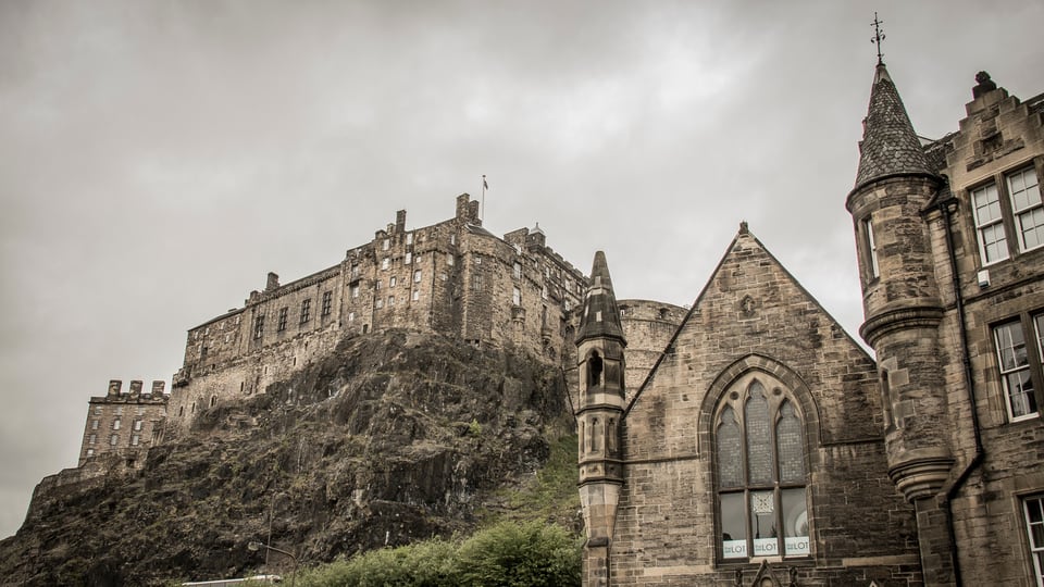 从 Grassmarket 仰望 Edinburgh Castle，深色城墙与岩壁压在 Old Town 石屋上方。