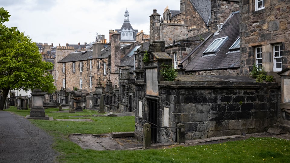Greyfriars Kirkyard 的墓碑与石砌纪念物沿草坡延伸，Edinburgh Old Town 屋顶围在后方。