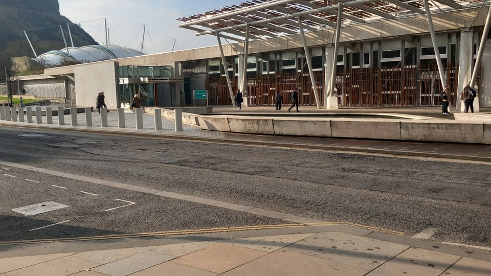Scottish Parliament 的钢木遮阳构件沿 Holyrood Road 展开，Arthur’s Seat 岩壁从建筑后方升起。