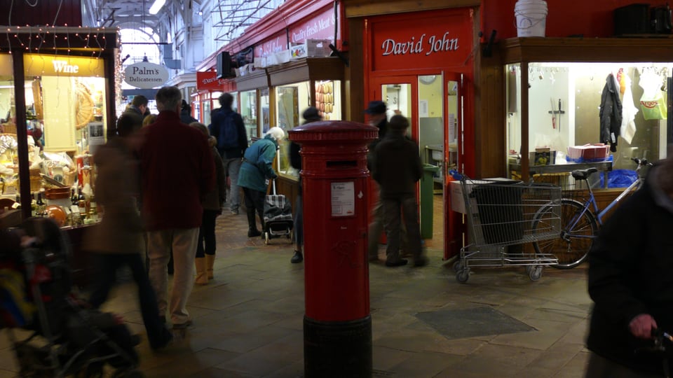 Oxford Covered Market 的室内通道由白色桁架覆盖，红色邮筒与相邻铺面构成市场街景。