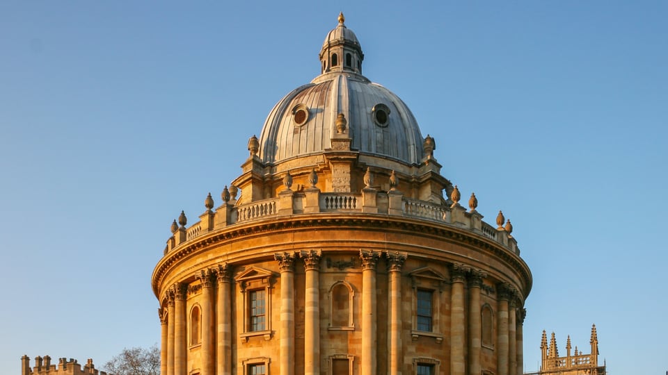 夕阳照亮 Radcliffe Camera 的圆形柱廊与铜色穹顶，周围是 Oxford 的石质学院建筑。